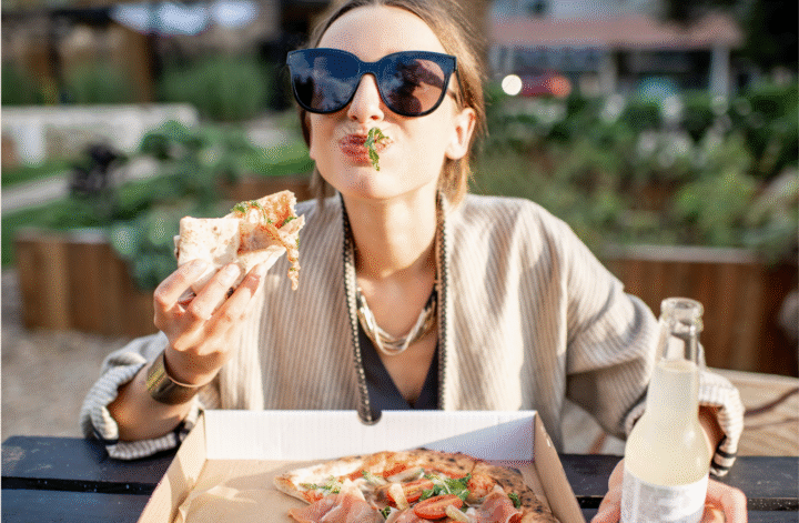 Frau mit Sonnenbrille isst entspannt ein Stück Pizza aus einem Karton – Symbolbild für intuitives Essen und bewussten Genuss im Alltag.